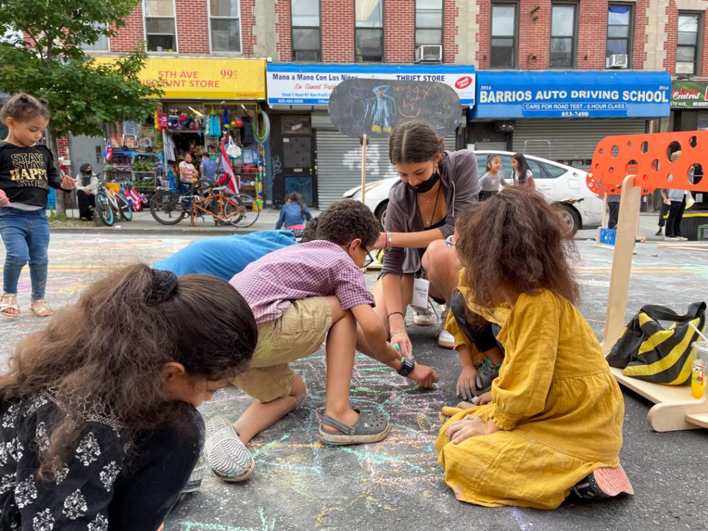 A group of young children and a woman are engaged in drawing with chalk on a city street. The background features colorful storefronts and bicycles parked nearby.