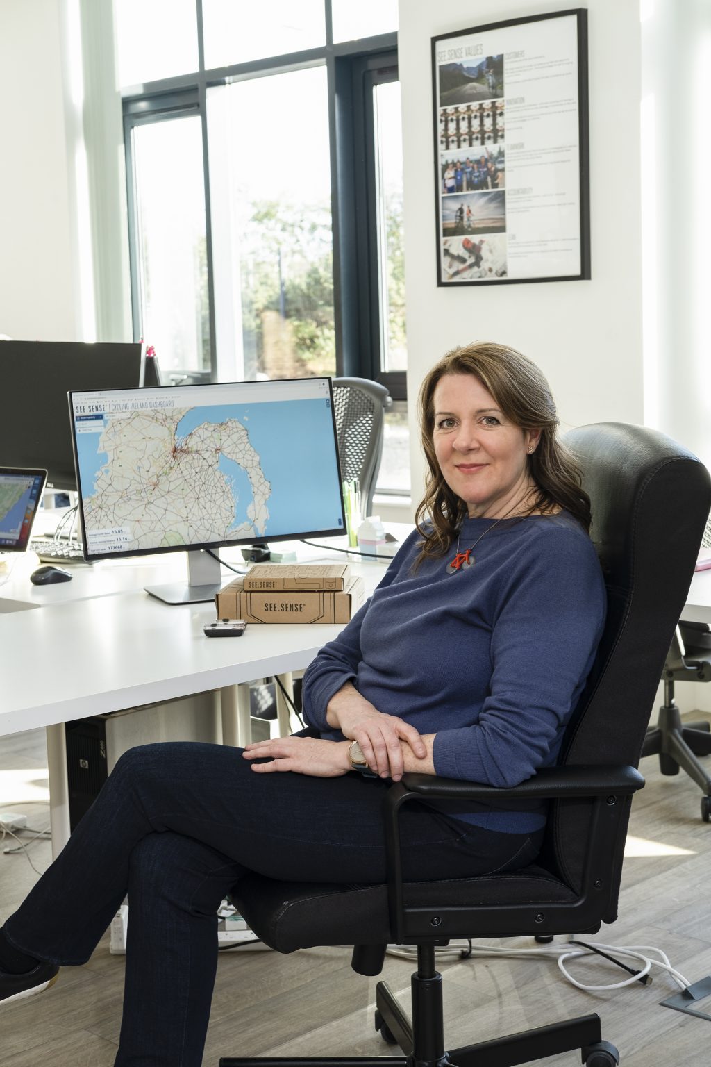 A woman in a blue sweater sits in an office chair, facing the camera with a slight smile. Behind her, a computer monitor displays a detailed map, and a poster with company values is visible on the wall.