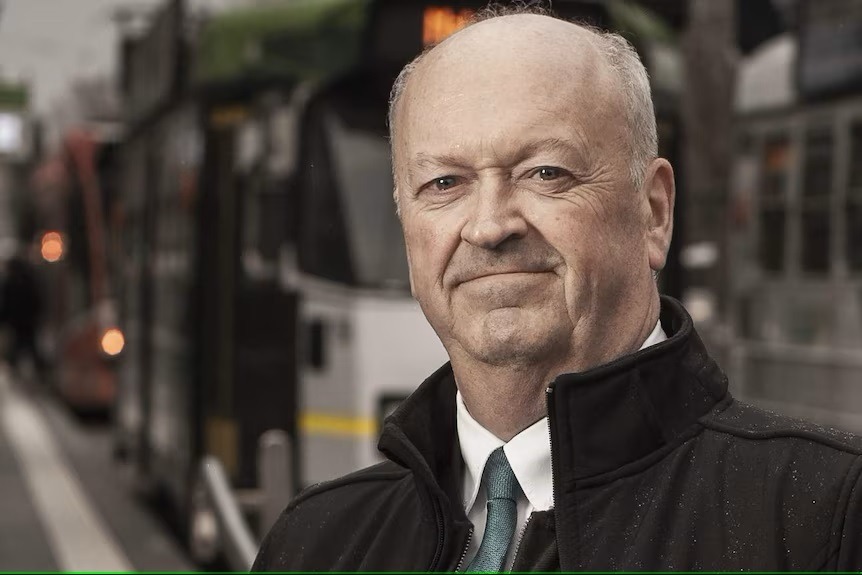 A man in a dark coat and suit stands confidently in front of a public transport vehicle, possibly a tram, with a city backdrop.