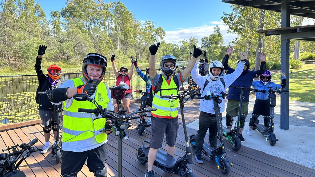 A group of people wearing helmets and high-visibility vests are gathered outdoors, riding electric scooters. They are smiling, raising their arms, and giving thumbs-up gestures. Trees and a pond are visible in the background, suggesting a recreational setting.