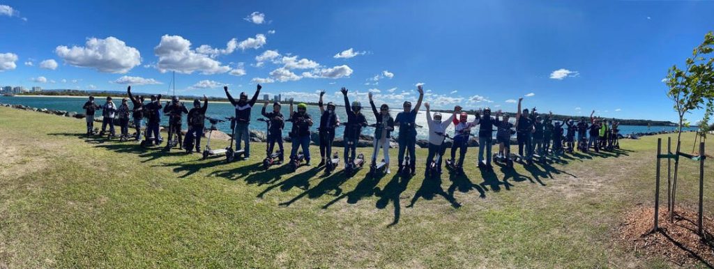 A group of people wearing helmets and protective gear stand in a line on a grassy area near the water, each holding an electric scooter. They raise their arms in celebration against a backdrop of blue sky, water, and distant city buildings.