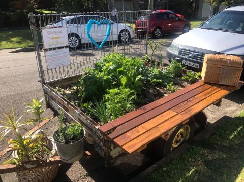 A box trailer parked on a suburban street, converted into a mobile garden with various plants, including leafy greens, growing in the trailer bed. The trailer features a wooden bench attached to its side and a wire fence around the garden. A blue heart decoration and a sign with text are visible on the fence.