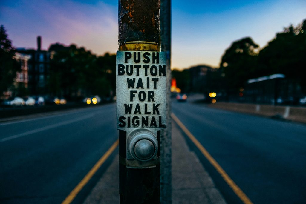 A pedestrian crosswalk button on a rusty pole with the instructions, "Push button wait for walk signal," in bold black letters. The background shows an empty road with blurred lights from distant cars as the evening sky fades from blue to purple.