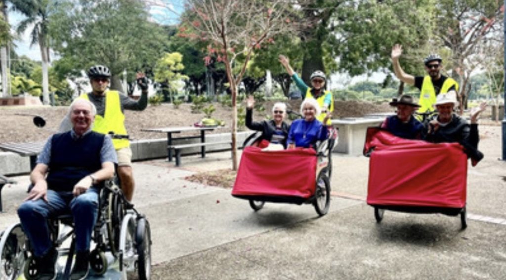 A group of smiling people outdoors enjoying a sunny day. Three trishaws with red covers are carrying elderly passengers, accompanied by volunteers wearing helmets and high-visibility vests. A man in a wheelchair sits nearby, also smiling. Trees and a picnic area can be seen in the background.