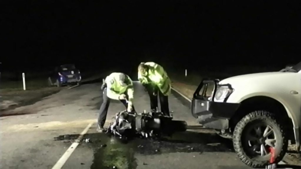 Two investigators in reflective jackets examine the wreckage of a motorcycle at night on a road. A white SUV is parked nearby, and a blue car is visible in the background.
