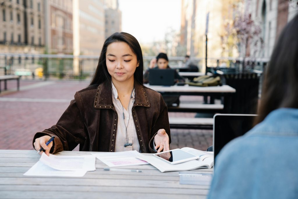 A woman with long black hair wearing a brown suede jacket sits at an outdoor table with papers, a tablet, and books spread out in front of her. She appears focused as she holds a pen and glances downward.