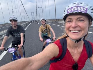 A smiling woman wearing a red top and a numbered cycling helmet takes a selfie while riding a bike on a large suspension bridge. Two teenage boys, also wearing helmets and riding bicycles, follow behind her. The bridge stretches into the distance under a partly cloudy sky.
