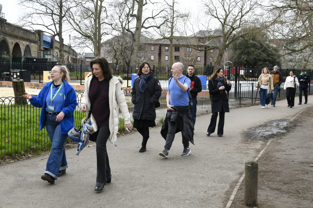 A group of people walking along a paved path in a park during daytime. Some are engaged in conversation, while others walk quietly. A woman in a blue uniform with a lanyard appears to be leading or guiding the group. The park has trees, a fenced playground, and a backdrop of brick residential buildings.
