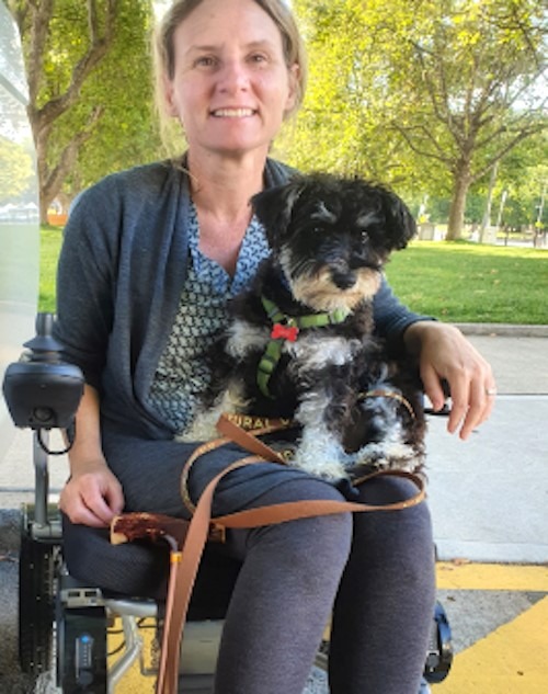 A woman with light skin, blonde hair, and a warm smile sits in a power wheelchair outdoors, holding a small black and white dog with a green harness and a leather leash on her lap. Trees and a grassy area are visible in the background.