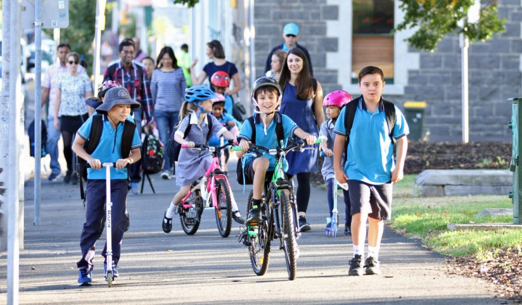 Kids biking to school, Children riding bikes, scooters, and walking to school along a tree-lined footpath, accompanied by parents and guardians.
