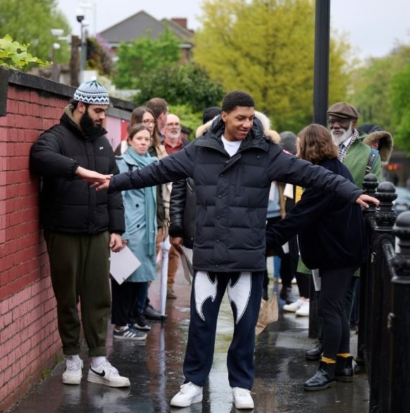 A vision-impaired man in a black puffer jacket uses outstretched arms to gauge the narrow width of a footpath, guided by touch along a red brick wall and metal railing. A diverse group stands behind him, observing.