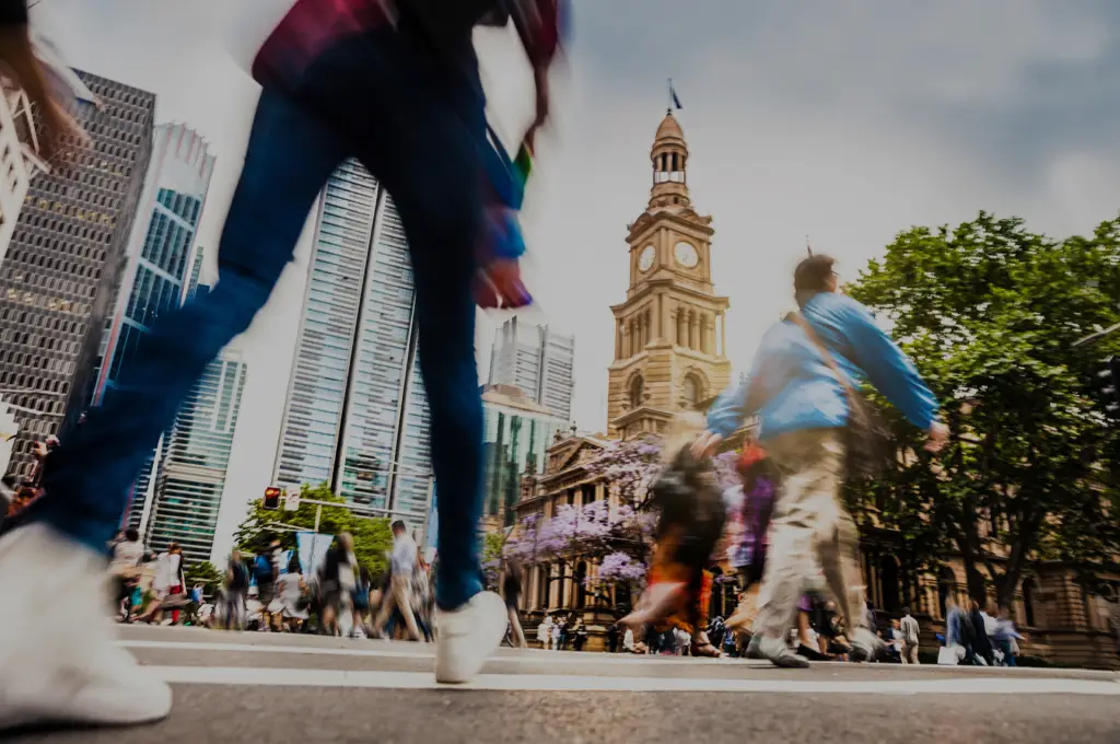 People walking across a busy urban intersection with tall buildings and a historic clock tower in the background.