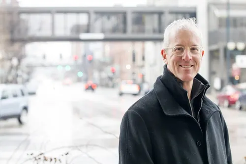 Jeff Speck, leading expert on walkability, smiling on a city street with traffic and a pedestrian bridge in the background.