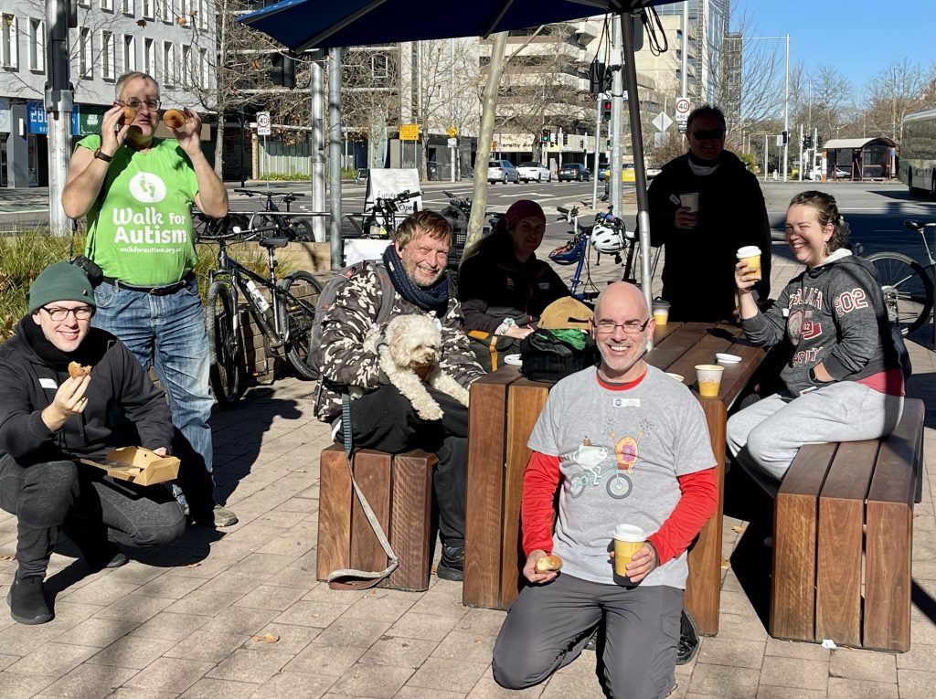 A group of cyclists, including Paris Lord, enjoying a break during a Canberra By Bike tour. They are sitting around a wooden table, with coffee cups and donuts, laughing and chatting. In the background, the urban environment of Canberra is visible, with bikes and pedestrians.