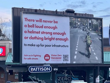 Large roadside billboard reading “There will never be a bell loud enough, a helmet strong enough or clothing bright enough to make up for poor infrastructure,” alongside an image of a cyclist riding in a protected bike lane at an intersection.