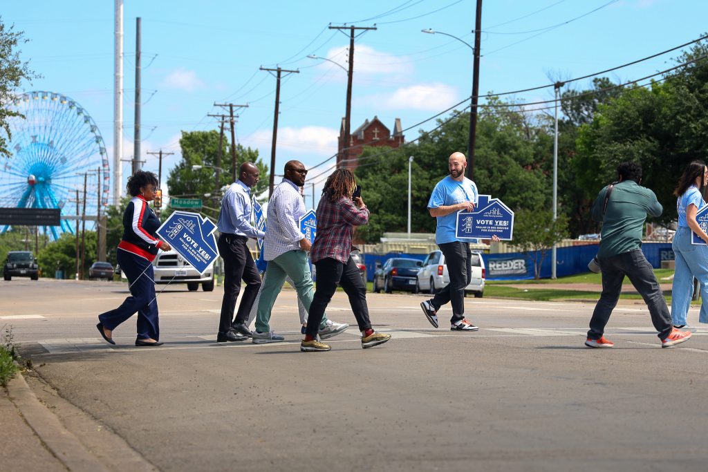 Community advocates walk together holding 'Vote Yes!' signs for Dallas housing reform, promoting affordable housing and parking policy changes.